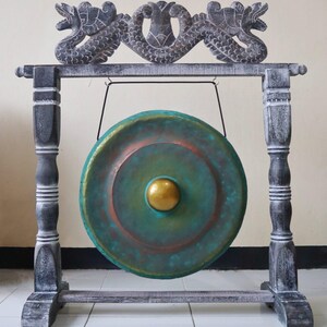 Puede incluir: Un gong decorativo con una cara de color turquesa y cobre y un centro dorado. El gong cuelga de un marco de madera con detalles de dragones tallados. El marco est&aacute; pintado en tonos grises y blancos.