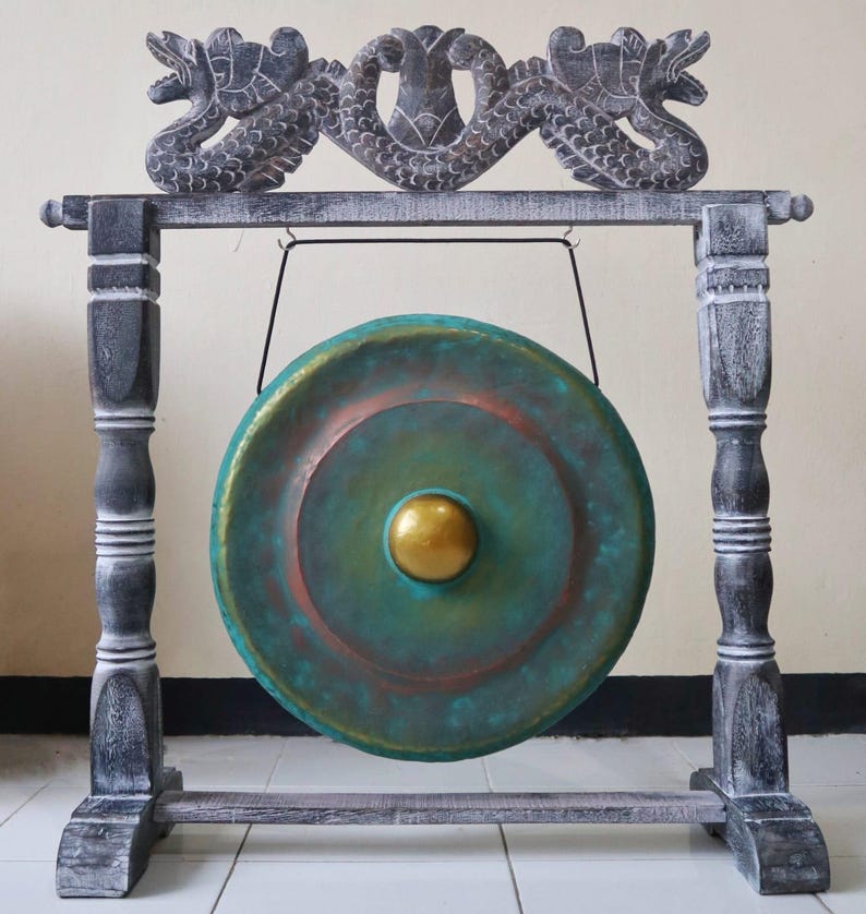 Puede incluir: Un gong decorativo con una cara de color turquesa y cobre y un centro dorado. El gong cuelga de un marco de madera con detalles de dragones tallados. El marco est&aacute; pintado en tonos grises y blancos.