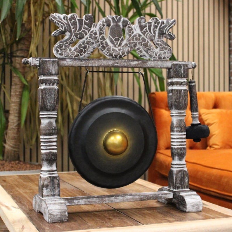 Puede incluir: Un gong decorativo con un disco de metal negro y un centro dorado, suspendido dentro de un marco de madera. El marco presenta figuras talladas en forma de drag&oacute;n y est&aacute; pintado en blanco y gris desgastado. Un mazo negro cuelga cerca.