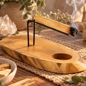 May include: A natural wood incense holder with a live edge, featuring a black metal stand that supports a burning palo santo stick. The base includes a circular indentation and a smooth, polished surface, showcasing the wood grain and texture.