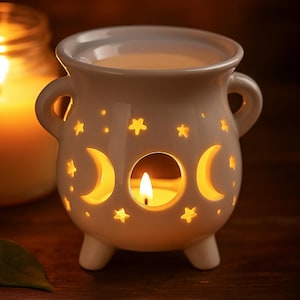 Puede incluir: Un portavelas de cerámica blanca con forma de caldero, con recortes de luna creciente y estrellas. El portavelas tiene dos asas y tres patas pequeñas. Una vela encendida está dentro, proyectando un brillo cálido.