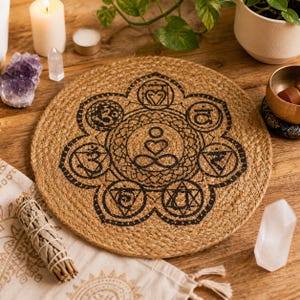 Puede incluir: Una esterilla de meditación redonda de yute tejido con un diseño de símbolo de chakra negro. La esterilla está rodeada de velas, cristales, un manojo de salvia y un pequeño cuenco tibetano, creando un ambiente sereno y espiritual.
