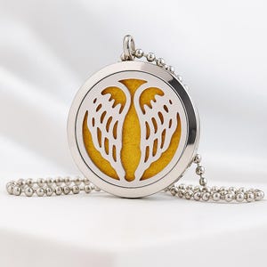 Puede incluir: Collar con colgante plateado con un diseño calado redondo con alas de ángel. El colgante tiene un interior amarillo y cuelga de una cadena de bolas. El collar se muestra sobre un fondo blanco.