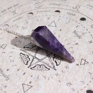 Può includere: Un pendolo di ametista sfaccettata con una catena d'argento, appoggiato su una superficie color crema con simboli astrologici e testo. Il pendolo è di colore viola scuro e termina a punta.