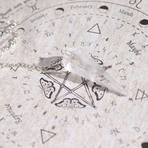Könnte beinhalten: Ein klarer Quarz-Kristall-Pendel mit einer silbernen Kette, auf einer cremefarbenen Oberfläche mit astrologischen Symbolen und Text. Das facettierte Pendel ist für Wahrsagerei und spirituelle Praktiken konzipiert.