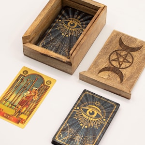 Könnte beinhalten: Holzkiste für Tarotkarten mit einem Kartenspiel mit einem Augendesign. Die Box ist geöffnet und zeigt die Karten im Inneren. Ein separater Holzdeckel mit Mond- und Sternendesign befindet sich neben der Box. Eine Tarotkarte mit dem Wort "WANDS" ist ebenfalls sichtbar.