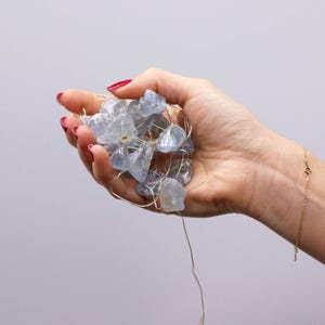 Può includere: Un pugno di cristalli azzurri, di forma irregolare, infilati su un sottile filo d'argento. I cristalli sono tenuti in una mano con smalto rosso, e un braccialetto d'oro è visibile sul polso. Lo sfondo è grigio chiaro.