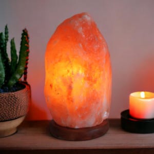 Könnte beinhalten: Eine leuchtend orangefarbene Himalaya-Salzlampe mit Holzsockel. Daneben ein kleiner Kaktus in einem dekorativen Topf und eine brennende Kerze in einem schwarzen Halter. Die Lampe strahlt ein warmes Licht aus.