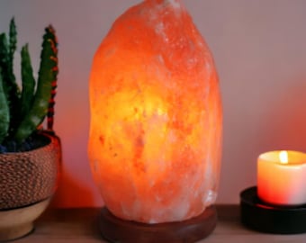 Könnte beinhalten: Eine leuchtend orangefarbene Himalaya-Salzlampe mit Holzsockel. Daneben ein kleiner Kaktus in einem dekorativen Topf und eine brennende Kerze in einem schwarzen Halter. Die Lampe strahlt ein warmes Licht aus.