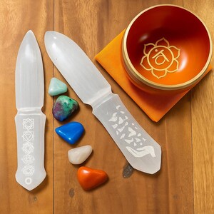 Könnte beinhalten: Ein Selenit-Zauberstab-Set mit einer Klangschale und farbigen Trommelsteinen. Die Zauberstäbe haben geätzte Designs und die Schale ist orange mit einem goldenen Blumendesign. Die Steine sind blau, grün, weiß und orange.