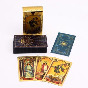 Puede incluir: Una caja y baraja de cartas del tarot doradas con un diseño celestial. La caja presenta una imagen de una figura, y la baraja incluye cartas con ilustraciones y títulos como "La Suma Sacerdotisa" y "El Loco". También hay un folleto azul.