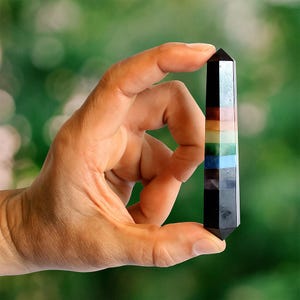 Op de afbeelding: Een veelkleurige kristallen obelisk, vastgehouden tussen vingers, heeft kleurstroken, waaronder zwart, bruin, rood, roze, geel, groen, blauw en paars. Het kristal is een rechthoekig prisma met een puntige bovenkant. De achtergrond is een vage groene kleur.