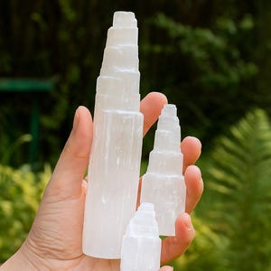 Può includere: Una mano tiene quattro torri di cristallo di selenite bianche di varie dimensioni. Le torri hanno un design a strati e a gradini, con un aspetto liscio e traslucido. Lo sfondo è un fogliame verde sfocato.