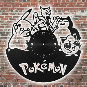 Può includere: Orologio in vinile nero con personaggi Pokemon, tra cui Pikachu, Meowth e Jigglypuff. Il quadrante dell'orologio ha uno sfondo bianco con numeri e lancette neri. La parola "Pokemon" è scritta in bianco sotto il quadrante dell'orologio.
