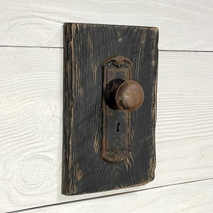May include: A rustic door knob and plate are mounted on a dark stained wooden plank. The knob is round and aged, with a keyhole below. The wood has a rough, textured appearance, adding to the vintage aesthetic.