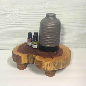 May include: A gray, ribbed, cylindrical essential oil diffuser with a black base, positioned next to two small amber glass bottles on a natural wood slab stand featuring a live edge and three short, cylindrical wooden legs.