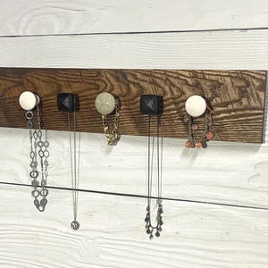May include: A wooden jewelry organizer with five decorative knobs. The dark brown wood board is mounted on a white wooden wall. Various necklaces and bracelets hang from the knobs, showcasing the organizer's functionality.