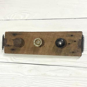 May include: A rustic wooden coat rack with three vintage door knobs. The knobs are brown, gold, and black, mounted on a weathered, rectangular wooden board. Hinges are attached on each end.