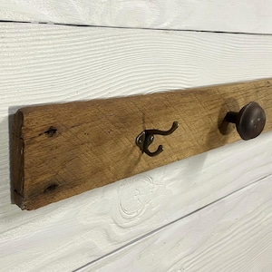 May include: A rustic wooden coat rack with two hooks and a knob. The wooden plank is a warm brown color, with visible wood grain and knots. The hooks are a dark bronze color, and the knob is a matching dark bronze. The rack is mounted on a white wooden wall.