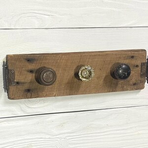 May include: A rustic wooden coat rack with three decorative knobs. The knobs are in varying styles: a bronze, a crystal, and a black knob. The rack is mounted on a white wooden wall.