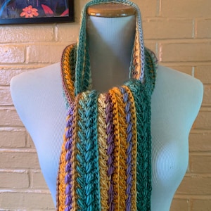 May include: A crochet scarf featuring vertical stripes in shades of teal, mustard yellow, and lavender. The scarf is constructed with a textured, raised stitch pattern that runs the length of the fabric, creating a ribbed appearance throughout the design.