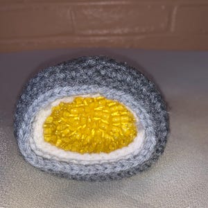 May include: Handmade crochet sushi roll with a gray, white, and yellow design. The outer layer is gray, followed by a white layer, and the center is filled with yellow beads, resembling the sushi's filling. A unique, handcrafted item.