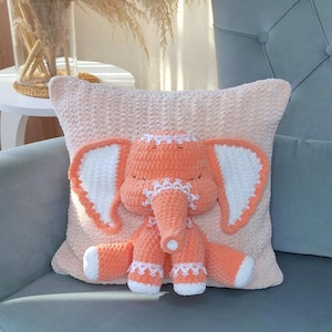 May include: A peach-colored crocheted elephant pillow with white crocheted ears and a white crocheted lace trim. The pillow is sitting on a light blue chair.