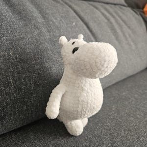 May include: White plush hippopotamus toy with black eyes. The toy is sitting on a gray couch.