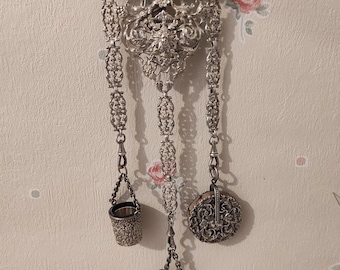 May include: An ornate silver-toned chatelaine with a decorative top and dangling accessories. The accessories include a small bucket, a round case, and a scissors holder. The chatelaine is suspended against a floral wallpaper background.