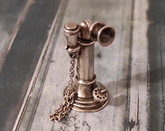 May include: A miniature silver-colored vintage telephone charm. The charm features a detailed design with a receiver, dial, and a chain. The telephone is approximately 1 inch tall, showcasing a classic, antique aesthetic.
