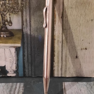 Może przedstawiać: Srebrny ołówek mechaniczny z teksturowanym korpusem i klipsami po bokach. Ołówek ma spiczasty grot i małą metalową pętlę na końcu.