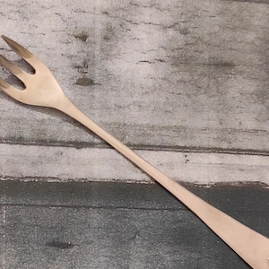 May include: A single, long-handled fork with three tines. The fork is made of a light-coloured metal, possibly silver or stainless steel, and has a simple, elegant design. The background is a weathered, grey-toned surface.