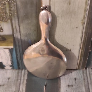 Puede incluir: Un espejo de mano plateado vintage con una cabeza en forma de lágrima y un mango largo y delgado. El espejo tiene una superficie reflectante y un pequeño lazo en la parte superior del mango. El espejo es probablemente de metal y es un artículo decorativo.