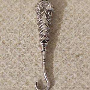 May include: A silver-toned metal button hook pendant featuring an ornate, textured handle with scrollwork details at the top. The slender shaft leads to a curved hook at the bottom, with small, stamped hallmark characters visible on the metal surface.