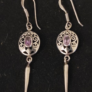 May include: A pair of silver drop earrings. Each earring has an oval pendant with an amethyst-coloured stone, encircled by an openwork pattern. A long, tapered silver element hangs below the pendant.