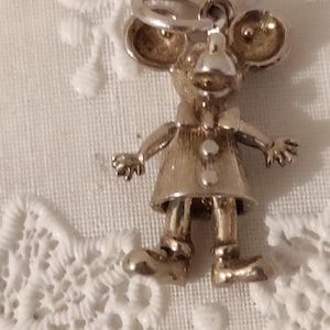 May include: A silver-toned charm in the shape of a mouse wearing a dress and bow tie. The mouse has large ears, a small nose, and is standing with its arms outstretched. The charm is likely for a bracelet or necklace.