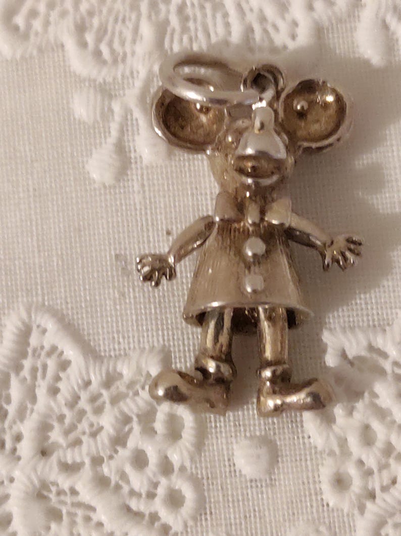 May include: A silver-toned charm in the shape of a mouse wearing a dress and bow tie. The mouse has large ears, a small nose, and is standing with its arms outstretched. The charm is likely for a bracelet or necklace.