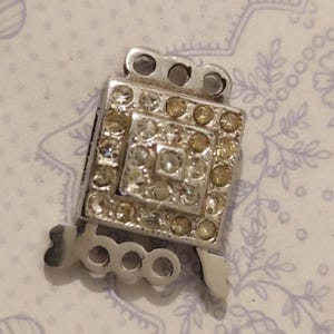 Pode incluir: Um fecho de joia prateado com um centro quadrado cravejado de strass. O quadrado é rodeado por strass menores, transparentes e âmbar. O fecho tem um design decorativo vazado.