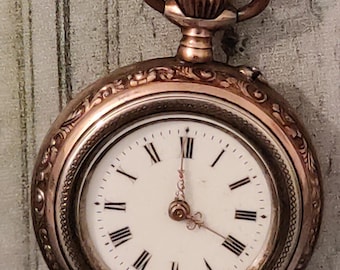 May include: A gold-toned pocket watch with a white face and black Roman numerals. The watch has an ornate, engraved design around the bezel and a loop at the top. The hands point to approximately 12:00. A classic timepiece.