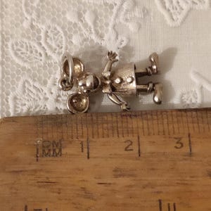 May include: A silver-toned charm in the shape of a mouse wearing a dress, lying on a wooden ruler. The charm has a detailed design with visible facial features, arms, and legs. The ruler shows inch measurements.