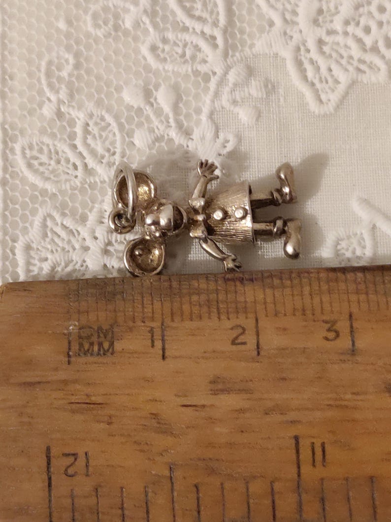 May include: A silver-toned charm in the shape of a mouse wearing a dress, lying on a wooden ruler. The charm has a detailed design with visible facial features, arms, and legs. The ruler shows inch measurements.