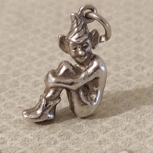 May include: A silver-toned metal charm shaped like a seated elf or pixie figure. The figure is depicted in a crouching position with knees pulled toward the chest, wearing a pointed hat and pointed shoes, with a small circular jump ring attached at the top.