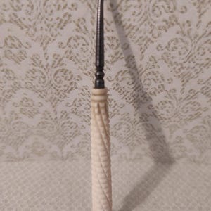 Op de afbeelding: Een knopenhaak met een donkere metalen schacht met een gebogen haak aan de bovenkant en een crèmekleurig handvat met een uitgesneden, spiraalvormige textuur die doet denken aan een touw. Het handvat eindigt in een kleine, ronde knop.