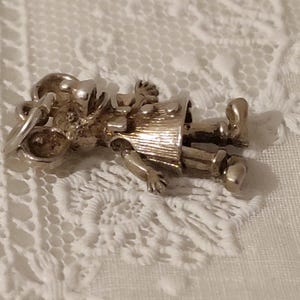 May include: A silver-toned charm in the shape of a small person, possibly a child, wearing a hat and a coat. The charm has articulated arms and legs. The charm is lying on a white lace surface.