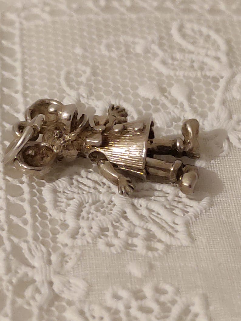 May include: A silver-toned charm in the shape of a small person, possibly a child, wearing a hat and a coat. The charm has articulated arms and legs. The charm is lying on a white lace surface.