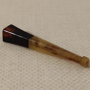 May include: A tapered, wooden pipe with a dark brown and black mouthpiece and a light brown body. The pipe has a small, cylindrical end. The wood grain is visible throughout the body of the pipe.
