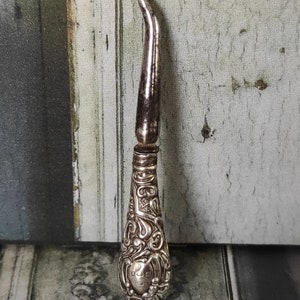 Peut inclure: Un chausse-pied en argent avec une poignée ornée et gravée. La poignée est en forme de larme et présente un motif floral.