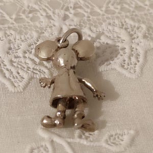 May include: A silver-colored charm in the shape of a cartoon mouse. The charm has large round ears, a dress, and small arms and legs. The charm is suspended from a small loop.