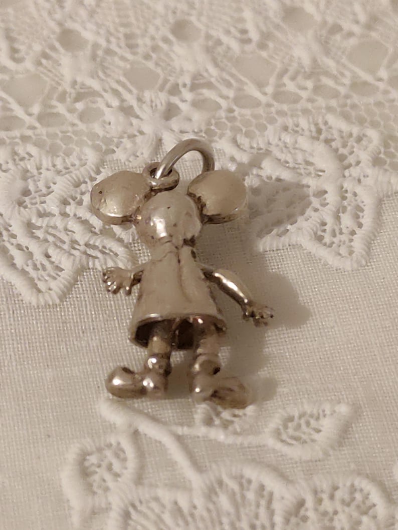 May include: A silver-colored charm in the shape of a cartoon mouse. The charm has large round ears, a dress, and small arms and legs. The charm is suspended from a small loop.