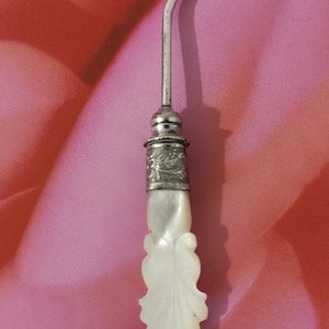 May include: An antique silver-toned button hook with a curved hook end and a decorative handle. The handle is made of white, carved material, possibly mother-of-pearl, with a leaf-like design. The hook is set against a pink background.